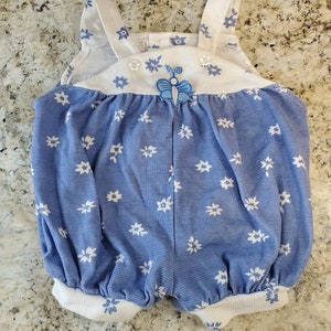 May include: A blue and white baby onesie with a white flower pattern and a butterfly applique. The onesie has white straps and a white trim around the legs.