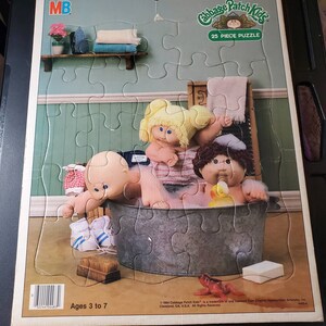May include: A 25-piece jigsaw puzzle featuring three Cabbage Patch Kids dolls in a bathtub. The dolls are wearing bathing suits and have blonde and brown hair. The puzzle is made for ages 3 to 7.