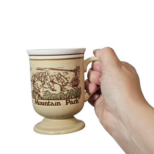 May include: A beige ceramic mug with a handle, featuring a brown illustration of people and buildings, with the text "Mountain Park". The mug has a pedestal base and a white rim.