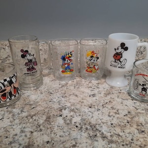 May include: Set of six vintage glass mugs featuring various Mickey Mouse characters. The mugs are clear glass with colorful cartoon images of Mickey Mouse, Minnie Mouse, and Steamboat Willie. The mugs are in good condition with no chips or cracks.