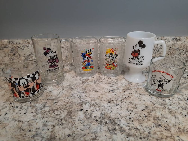 May include: Set of six vintage glass mugs featuring various Mickey Mouse characters. The mugs are clear glass with colorful cartoon images of Mickey Mouse, Minnie Mouse, and Steamboat Willie. The mugs are in good condition with no chips or cracks.
