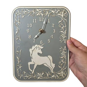 May include: A rectangular wall clock with a gray face, featuring a white unicorn silhouette at the bottom and white floral border detailing. The clock has white hour and minute hands, and white numbers from one to twelve in a serif font.
