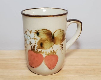 May include: A white ceramic mug with a brown rim and handle. The mug has a painted design of two red strawberries with green leaves and a white flower.