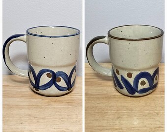 May include: Two white ceramic mugs with blue and brown designs. The mugs have a speckled glaze and a brown rim.