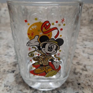 May include: A clear glass cup with a Mickey Mouse design. Mickey is wearing a safari outfit and holding binoculars. The cup has a red, yellow, and white confetti design.