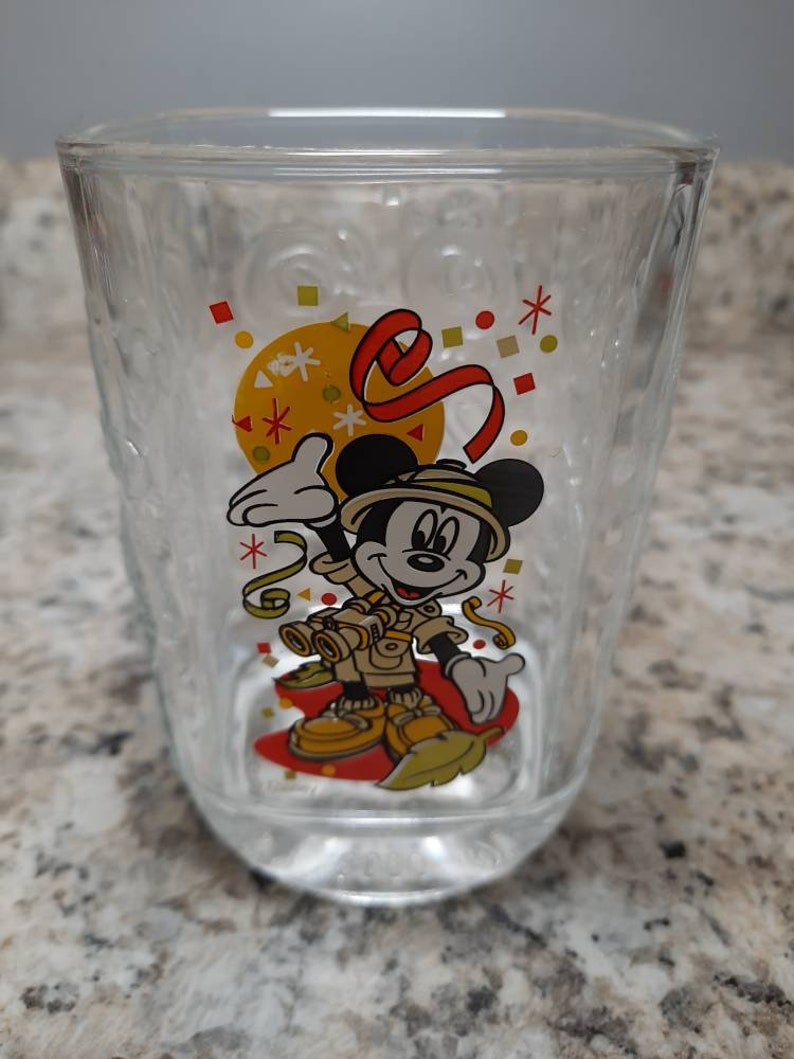 May include: A clear glass cup with a Mickey Mouse design. Mickey is wearing a safari outfit and holding binoculars. The cup has a red, yellow, and white confetti design.