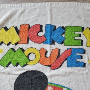 May include: White beach towel with a colorful Mickey Mouse design. The words "Mickey Mouse" are in a bold, colorful font with a yellow, green, blue, red, and orange color scheme. There are small red stars scattered around the design.