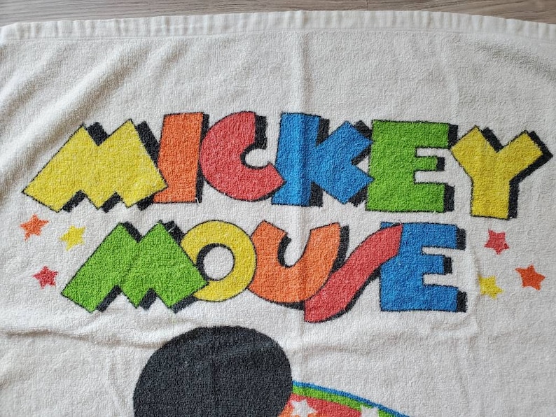 May include: White beach towel with a colorful Mickey Mouse design. The words "Mickey Mouse" are in a bold, colorful font with a yellow, green, blue, red, and orange color scheme. There are small red stars scattered around the design.