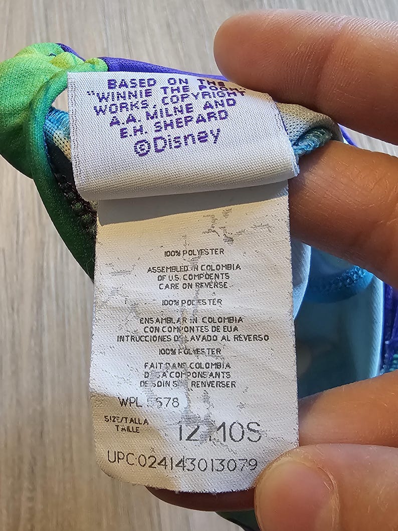 May include: A tag from a Winnie the Pooh garment, featuring text including "&copy;Disney", "100% POLYESTER", and size "12 10S". The tag is white with black text and is attached to a colorful fabric.