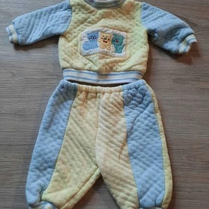 May include: A two-piece quilted outfit consisting of a long-sleeved pullover and matching pants. The set features a pale yellow and light blue color scheme with a central graphic patch on the chest depicting three small cartoon animals in blue, yellow, and green.