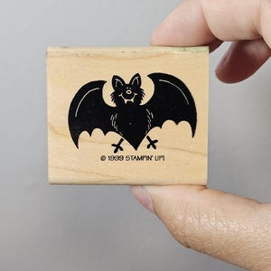 Pode incluir: Um carimbo de madeira com um desenho de morcego preto com asas abertas. O morcego tem uma cara sorridente e est&aacute; sobre um bloco de madeira clara. O texto "&copy; 1999 STAMPIN' UP!" est&aacute; impresso abaixo.