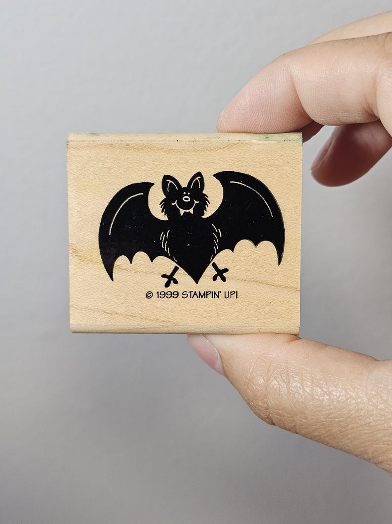 Pode incluir: Um carimbo de madeira com um desenho de morcego preto com asas abertas. O morcego tem uma cara sorridente e est&aacute; sobre um bloco de madeira clara. O texto "&copy; 1999 STAMPIN' UP!" est&aacute; impresso abaixo.