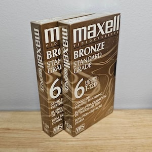 May include: Two Maxell VHS video cassette tapes in bronze color with the text "Maxell Video Cassette Bronze Standard Grade" and "6 Hours in EP Mode T-120" printed on the front. The tapes are labeled "VHS" and "T-120".
