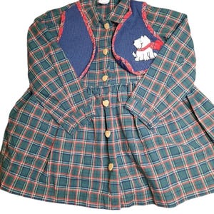 May include: A green, red, and white plaid dress with a blue vest and a white dog applique. The vest has a red trim and the dog has a red scarf.