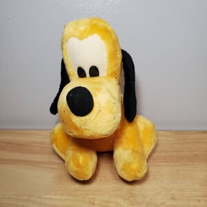 May include: A yellow plush toy of Pluto, a cartoon dog from Disney. The toy is sitting with its legs crossed and has black ears and a black nose.