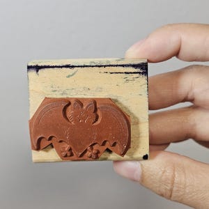 Pode incluir: Um carimbo de borracha castanho com um desenho de morcego. O morcego tem uma cara sorridente e est&aacute; montado num bloco de madeira retangular. O carimbo &eacute; segurado por uma m&atilde;o contra um fundo neutro. Ideal para trabalhos manuais de Halloween.