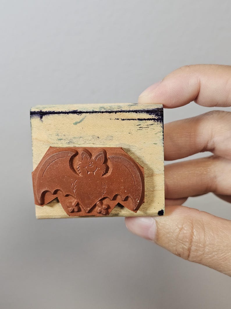 Pode incluir: Um carimbo de borracha castanho com um desenho de morcego. O morcego tem uma cara sorridente e est&aacute; montado num bloco de madeira retangular. O carimbo &eacute; segurado por uma m&atilde;o contra um fundo neutro. Ideal para trabalhos manuais de Halloween.
