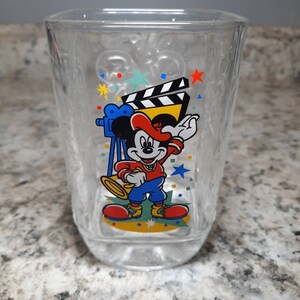 May include: A clear glass cup with a Mickey Mouse design. Mickey is wearing a red hat and holding a trumpet. The design also includes a film clapboard, a camera, and confetti.