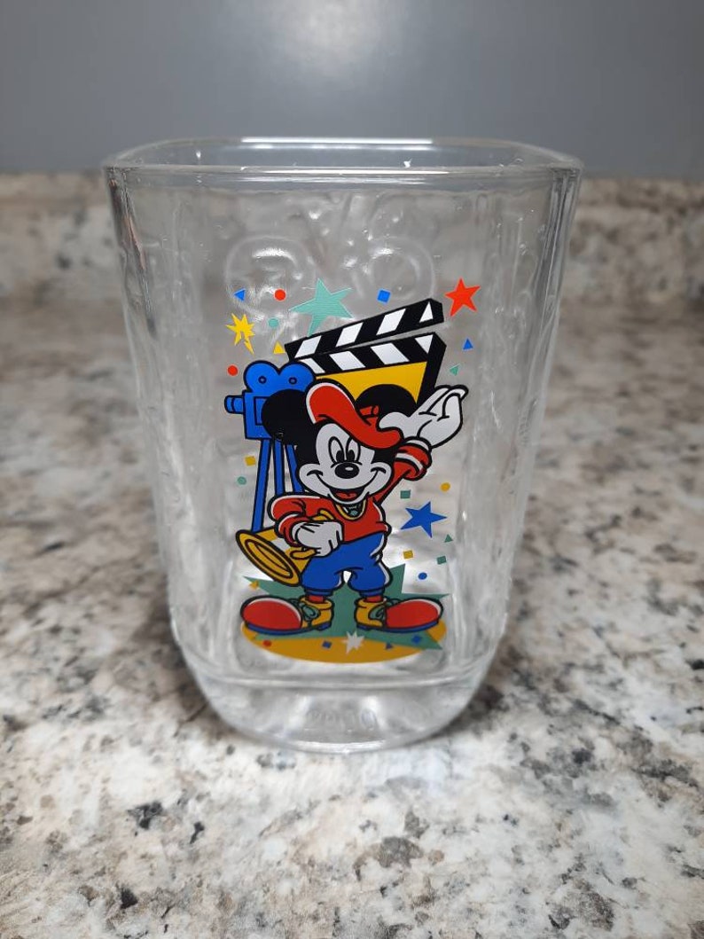 May include: A clear glass cup with a Mickey Mouse design. Mickey is wearing a red hat and holding a trumpet. The design also includes a film clapboard, a camera, and confetti.