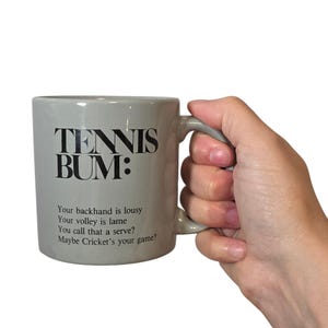 May include: A light gray ceramic mug featuring black text that reads TENNIS BUM: Your backhand is lousy, Your volley is lame, You call that a serve?, Maybe Cricket's your game? The mug has a curved handle on the right side and a smooth finish.