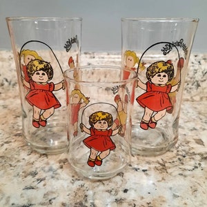 May include: Set of three clear glass drinking glasses with a colorful illustration of a girl skipping rope. The glasses feature the text "Cabbage Patch Kids".
