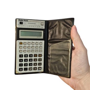 May include: A black and silver Radio Shack EC-4004 scientific calculator with a leather case. The calculator has a display screen, a keypad with numbers, functions, and operations, and a RUN button.