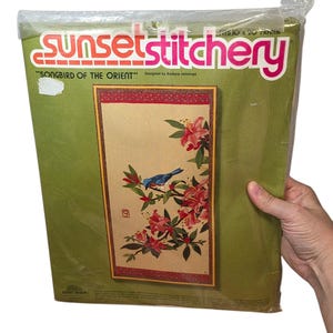 May include: A Sunset Stitchery embroidery kit titled Songbird of the Orient, designed by Barbara Jennings, packaged in a clear plastic bag. The kit features a blue bird on a branch with pink flowers, sized for a 10 inch by 20 inch frame.