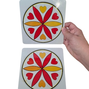May include: Two square white coasters featuring a circular mandala-style design. The pattern consists of four red petals, four yellow petals, and four red hearts arranged symmetrically within a black and yellow circular border on each coaster.