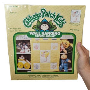 May include: A Cabbage Patch Kids wall hanging stenciling kit in a yellow box. The kit includes a stencil design, fabric, paint, and instructions. The finished size is approximately 22 x 23 inches. The box features a yellow and green design with the Cabbage Patch Kids logo.