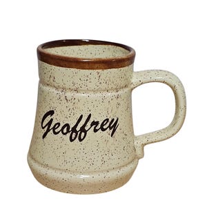 May include: Cream-colored ceramic mug with a speckled design and a brown rim and handle. The name "Geoffrey" is written in a black script on the front of the mug. The mug has a classic, sturdy shape.
