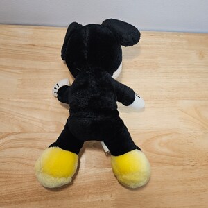 May include: Black plush toy with yellow shoes, resembling a cartoon character. The toy is sitting on a wooden surface.
