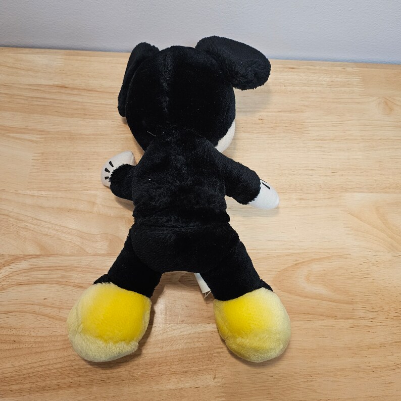 May include: Black plush toy with yellow shoes, resembling a cartoon character. The toy is sitting on a wooden surface.
