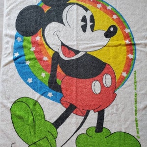 May include: A white beach towel with a colorful rainbow and stars surrounding a cartoon image of Mickey Mouse. Mickey Mouse is wearing red shorts and green shoes. The towel has the text "Walt Disney Productions" and "Printed in U.S.A."