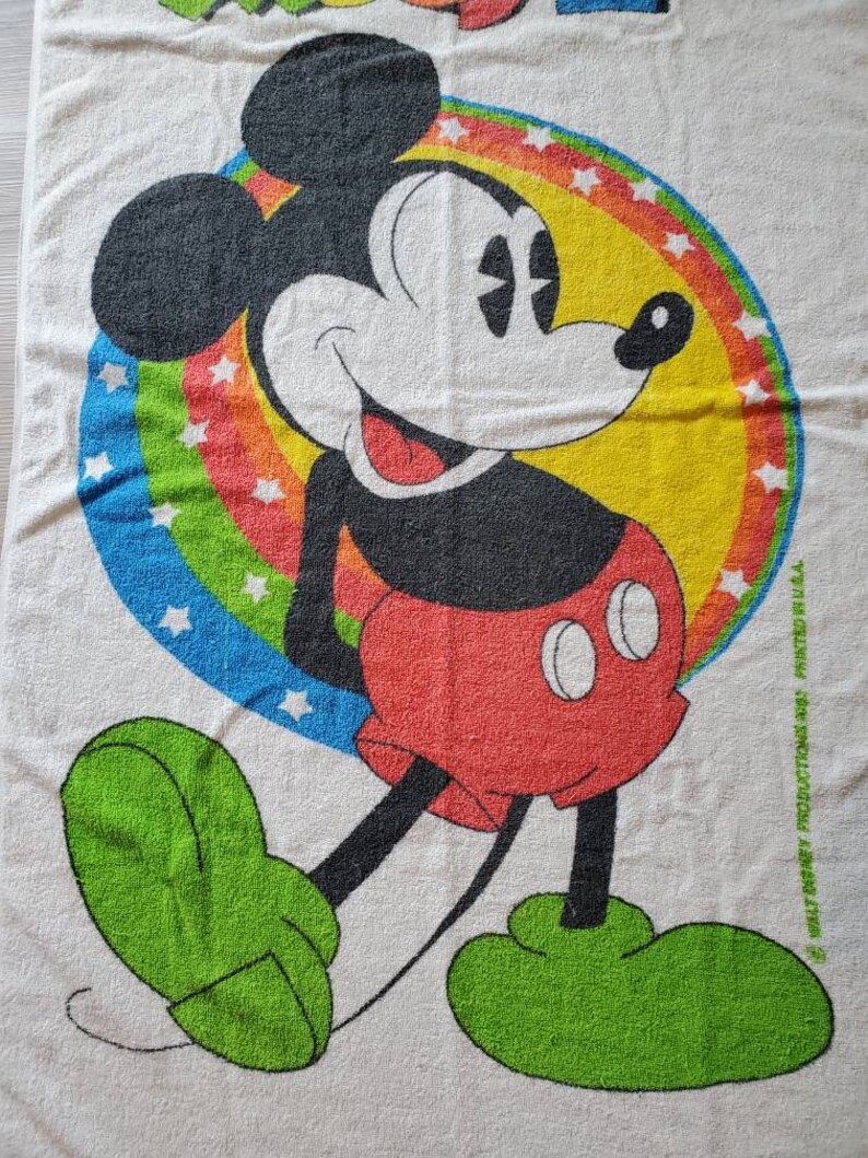 May include: A white beach towel with a colorful rainbow and stars surrounding a cartoon image of Mickey Mouse. Mickey Mouse is wearing red shorts and green shoes. The towel has the text "Walt Disney Productions" and "Printed in U.S.A."