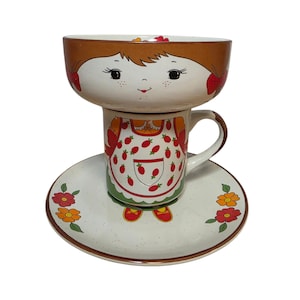 May include: A whimsical ceramic breakfast set featuring a bowl, mug, and saucer. The bowl and mug are designed with a smiling face and brown hair. The mug has a strawberry apron design, and the saucer is decorated with flowers.