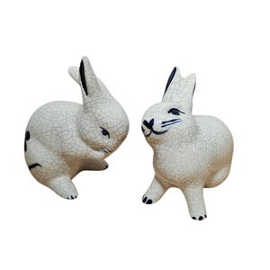 May include: Two ceramic rabbit figurines. The rabbits are white with a crackle glaze and black accents on the ears, eyes, and feet. One rabbit is posed looking down, and the other is looking forward.
