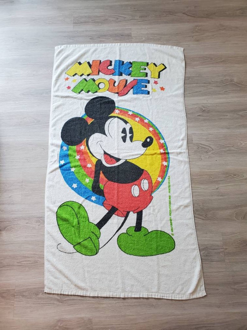 May include: White beach towel with a colorful Mickey Mouse design. The towel features Mickey Mouse walking with a red and yellow circle around him. The text "Mickey Mouse" is printed in rainbow colors at the top of the towel.