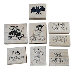 Pode incluir: Uma cole&ccedil;&atilde;o de carimbos de madeira com tema de Halloween. Os desenhos incluem um morcego, uma bruxa numa vassoura, um gato numa l&aacute;pide, um esqueleto e frases como "Happy Halloween!" e "Best Witches". Cada carimbo tem um desenho em tinta preta.