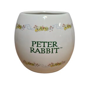 May include: White ceramic Peter Rabbit mug with a rounded shape. The mug features the text "PETER RABBIT" in green, with floral and vine accents in yellow, pink, and blue. The mug is a collectible item.