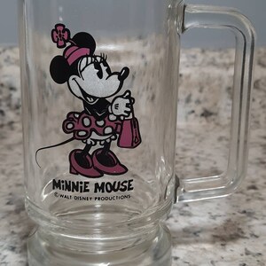 May include: A clear glass mug with a handle features a cartoon image of Minnie Mouse in a pink dress with white polka dots. The mug has the text "Minnie Mouse" and "&copy; WALT DISNEY PRODUCTIONS" printed on the bottom.