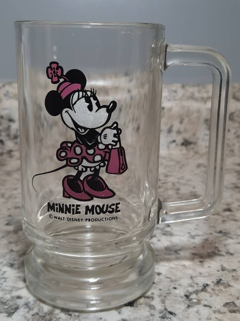 May include: A clear glass mug with a handle features a cartoon image of Minnie Mouse in a pink dress with white polka dots. The mug has the text "Minnie Mouse" and "&copy; WALT DISNEY PRODUCTIONS" printed on the bottom.