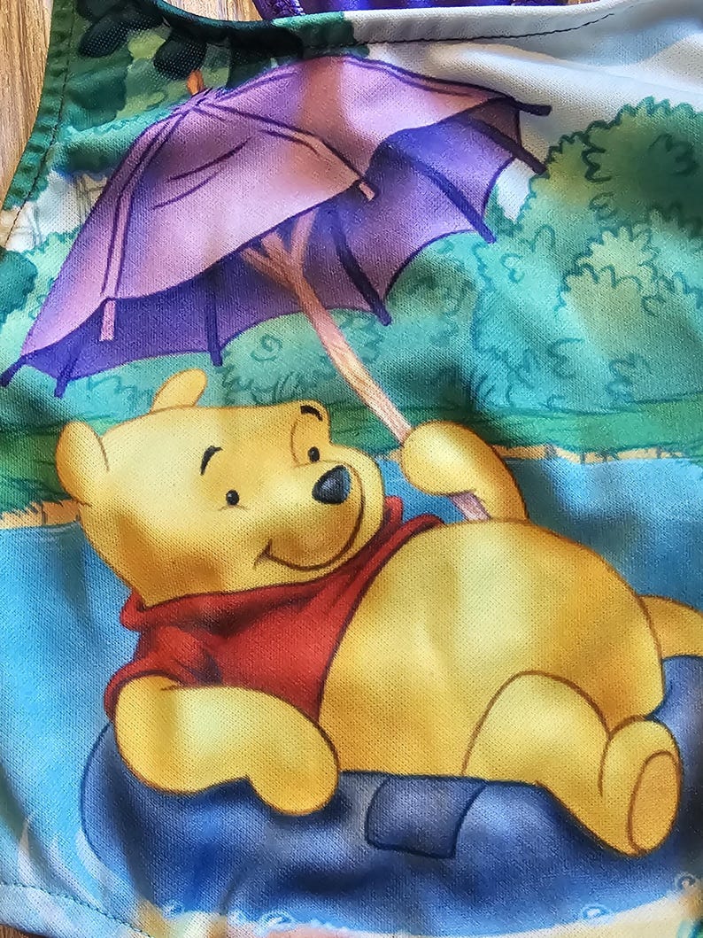 May include: A yellow cartoon bear wearing a red shirt, holding a purple umbrella, and resting on a blue inflatable ring. The background features a blue and green landscape. The image is from a children's product.