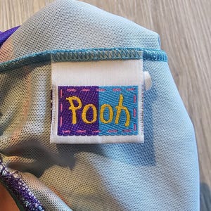 May include: Close-up of a light blue fabric with a sewn-on label. The label is divided into purple and blue sections, with the word "Pooh" embroidered in yellow and pink thread. The fabric has a textured appearance.