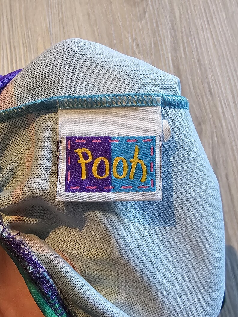 May include: Close-up of a light blue fabric with a sewn-on label. The label is divided into purple and blue sections, with the word "Pooh" embroidered in yellow and pink thread. The fabric has a textured appearance.