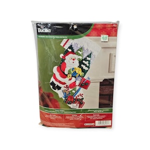 May include: A Bucilla Christmas stocking kit in a clear plastic package. The stocking features a felt design of Santa Claus, gifts, and Christmas trees. The stocking measures 18 inches (45.72 cm) in length.