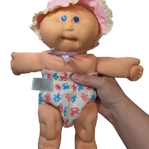 May include: A doll with a soft peach-colored body, blue eyes, and a round face. The doll wears a pink bonnet with white lace trim and a light-colored diaper featuring a repeating pattern of small pink and blue rocking horses and a blank tag.