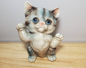 May include: A small ceramic figurine of a gray and white kitten with blue eyes. The kitten is sitting up with its paws raised, as if it is playing.