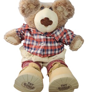 May include: A brown plush bear wearing a red and blue plaid shirt, beige pants, and brown boots with the words "FurSkins" on the soles.