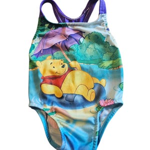May include: One-piece swimsuit featuring a colorful illustration of Winnie the Pooh relaxing in a pond. The swimsuit has a blue and purple color scheme with a cartoon scene of Pooh Bear under an umbrella. The straps are purple.