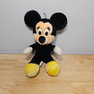 May include: A black and yellow plush toy of Mickey Mouse's girlfriend Minnie Mouse. She has large black ears, a black nose, and yellow shoes.
